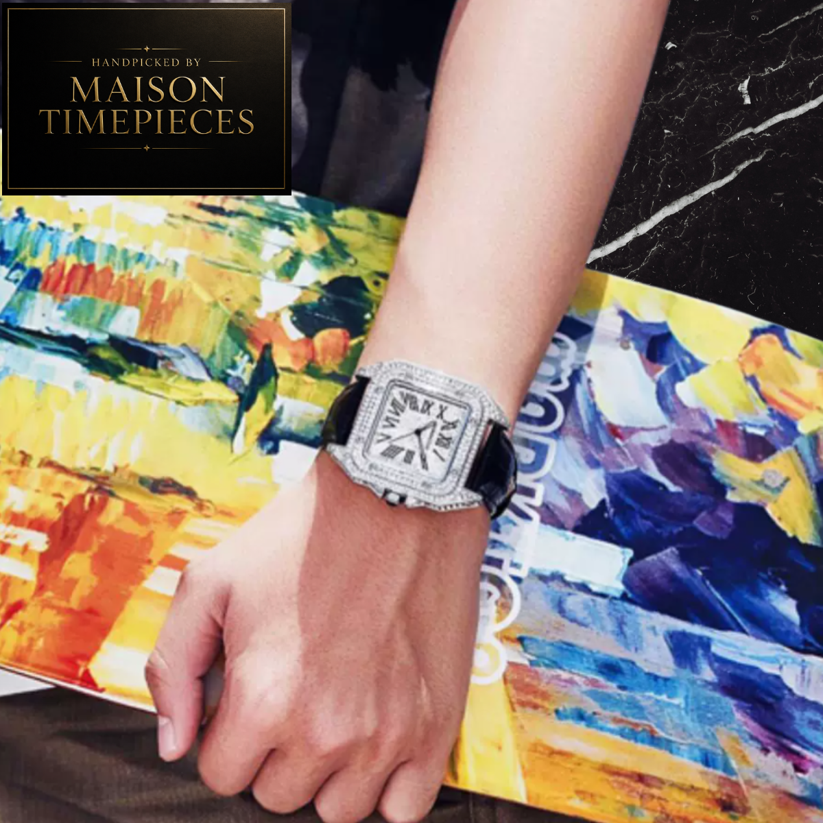 A close-up of a person's hand and wrist wearing a Maison Timepieces watch with a square silver-toned case encrusted with clear stones and a black strap, resting on a colorful abstract painting.