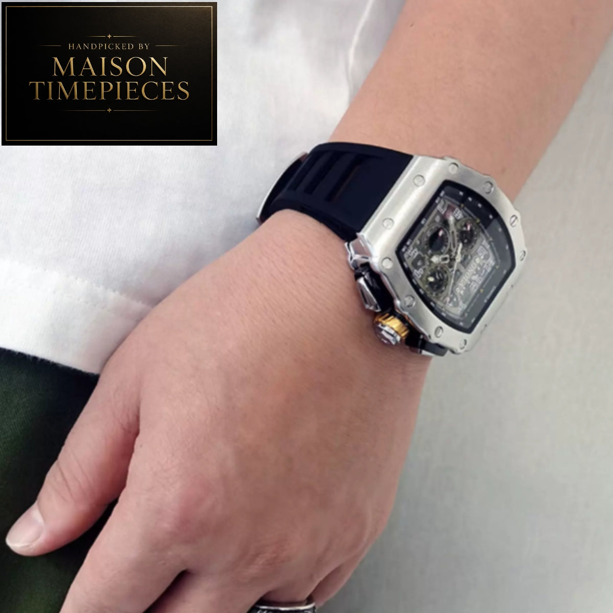 A close-up of a wrist wearing a Maison Timepieces skeleton watch with a silver-toned case, black dial, and black rubber strap. A Maison Timepieces logo badge is visible in the top-left corner.
