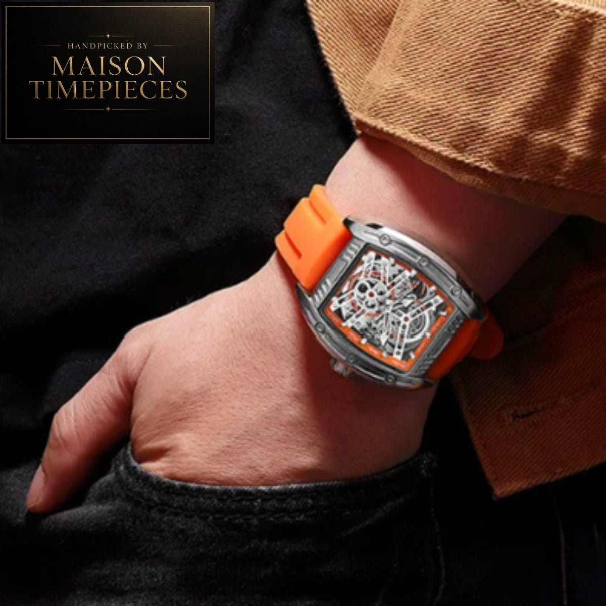 A close-up of a wrist wearing a Maison Timepieces watch with a bright orange rubber strap and a silver-toned case showing a skeleton dial. A hand rests in a pocket beside it, with a brown jacket cuff visible, and a Maison Timepieces logo badge appears in the top-left corner.