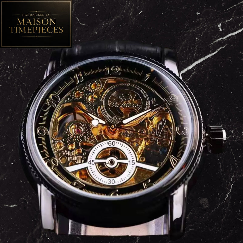 A Maison Timepieces skeleton watch with a black leather strap, featuring a transparent crystal case that reveals intricate gold-toned mechanical gears and a white subdial at the bottom, set against a dark marble background.