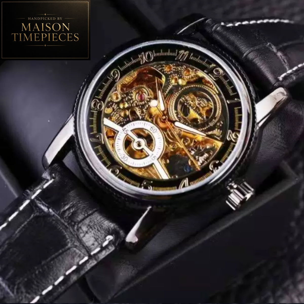A close-up of a skeleton watch with a black leather strap, silver case, and highly visible gold-toned mechanical movement inside a black presentation box. A logo in the top-left corner reads 'Handpicked by Maison Timepieces'.
