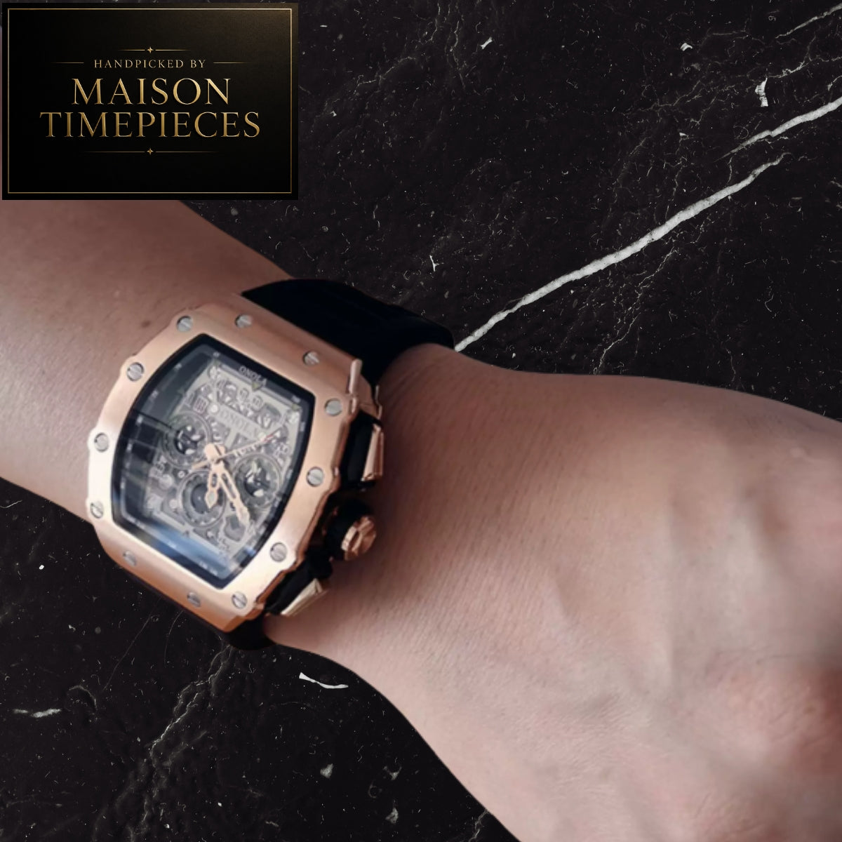 A luxury wristwatch with a rose gold-toned case, skeleton dial, and black strap, shown worn on a person's wrist against a dark marble background. A 'Handmade by Maison Timepieces' logo is visible in the top-left corner.