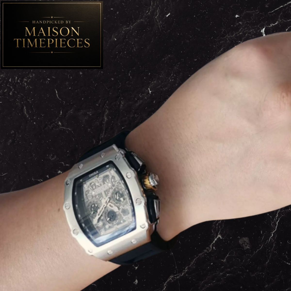 A close-up of a wrist wearing a Maison Timepieces men's watch with a square silver-toned case, black dial, and black strap, set against a dark marble background with a Maison Timepieces logo in the top-left corner.