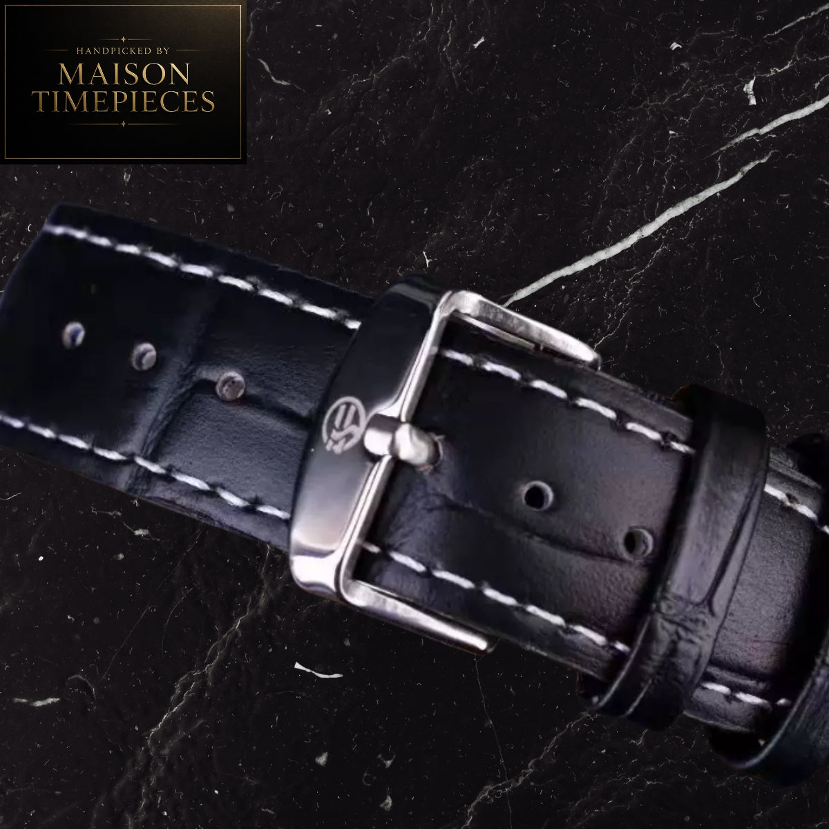 A close-up of a black leather watch strap with white contrast stitching and a silver-toned buckle, displayed on a dark marble surface. A Maison Timepieces logo badge is visible in the top-left corner.