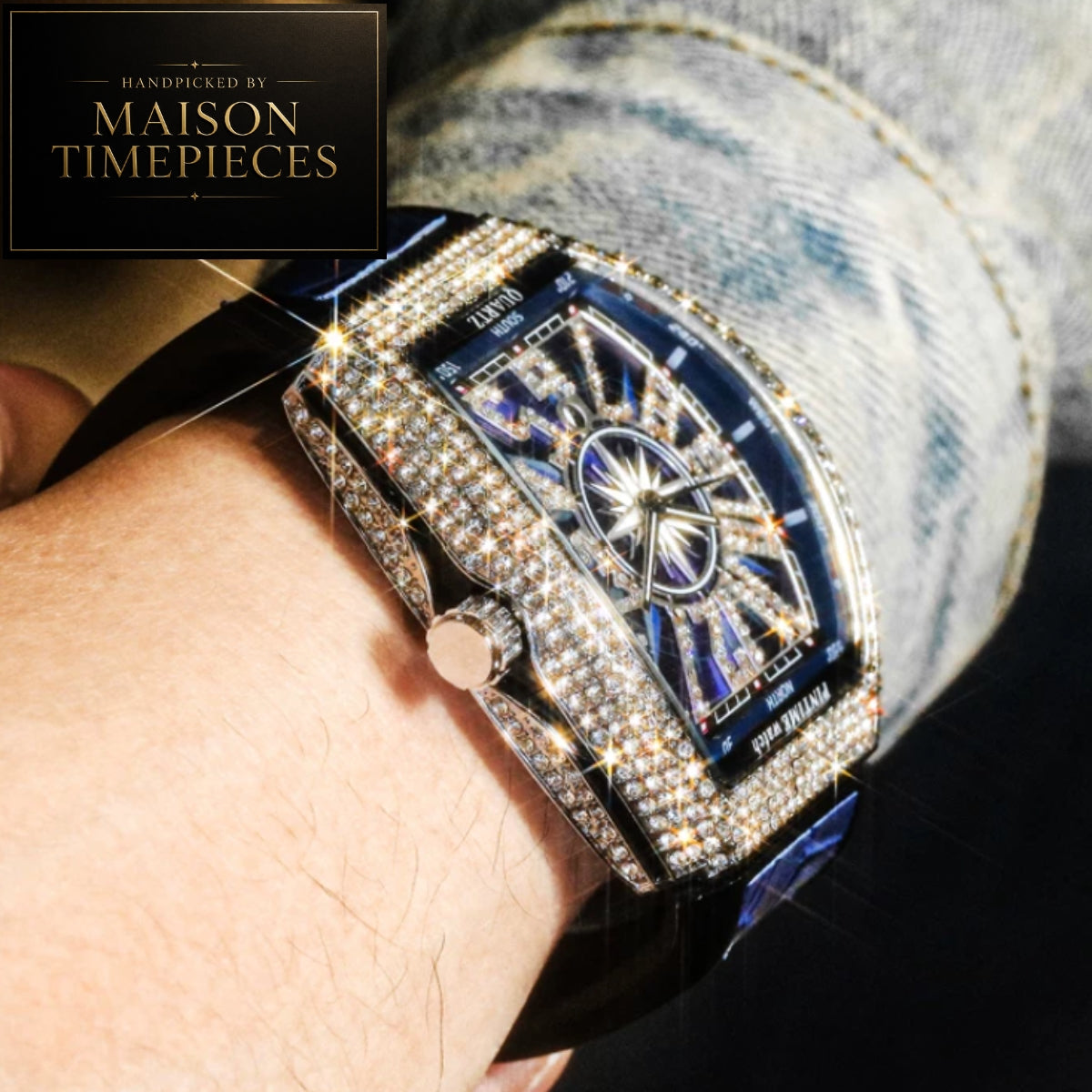 A close-up lifestyle shot of a wrist wearing a highly embellished watch. The watch features a rectangular, diamond-encrusted case, a dark blue dial with sunburst detailing, and a black strap.