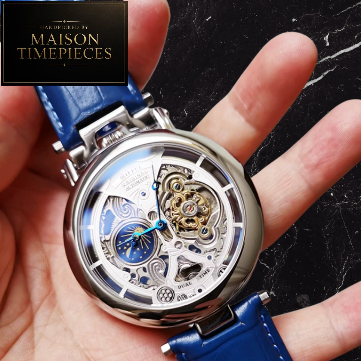A close-up of a skeleton watch with a blue leather strap held in a hand. The watch has a silver-toned case, a transparent dial showing intricate gold and silver mechanical gears, and a blue moonphase subdial.