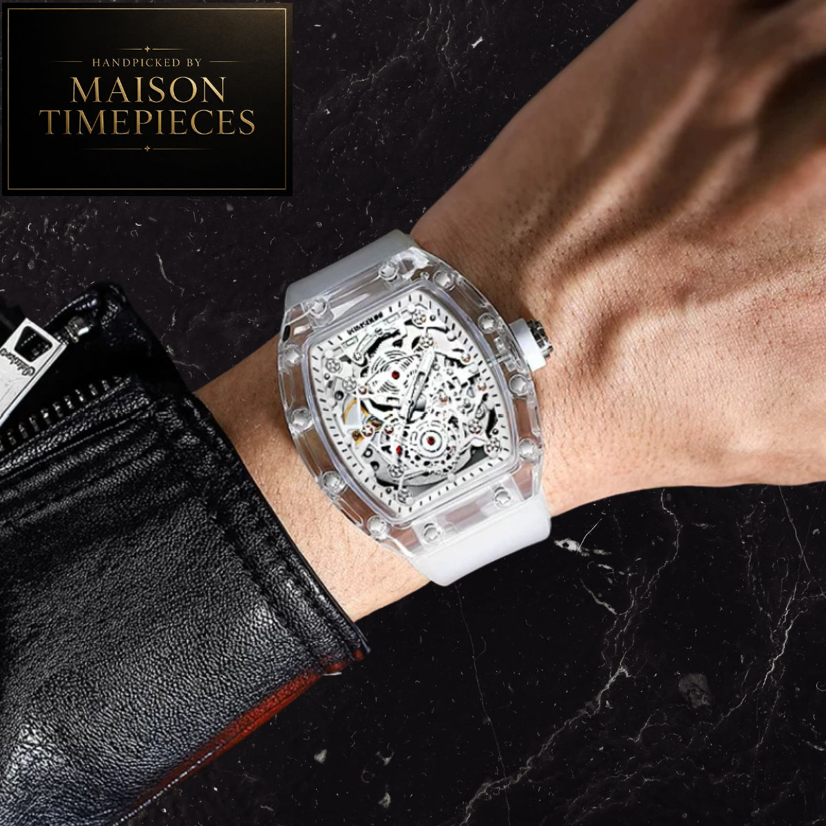 A skeleton watch with a clear case and white strap worn on a wrist, shown against a black marble background with a black leather sleeve. A logo in the top left reads 'Handcrafted by Maison Timepieces'.