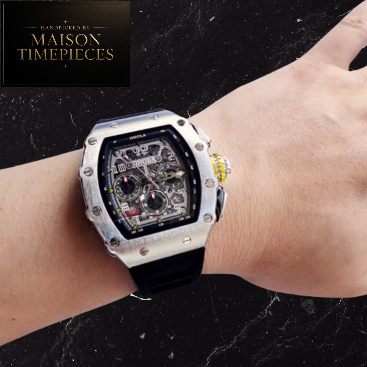 A silver-tone Richard Mille-style wristwatch with a black strap and skeleton dial, shown worn on a person's wrist against a dark marble background. A black logo badge in the top-left reads 'Handcrafted by Maison Timepieces'.