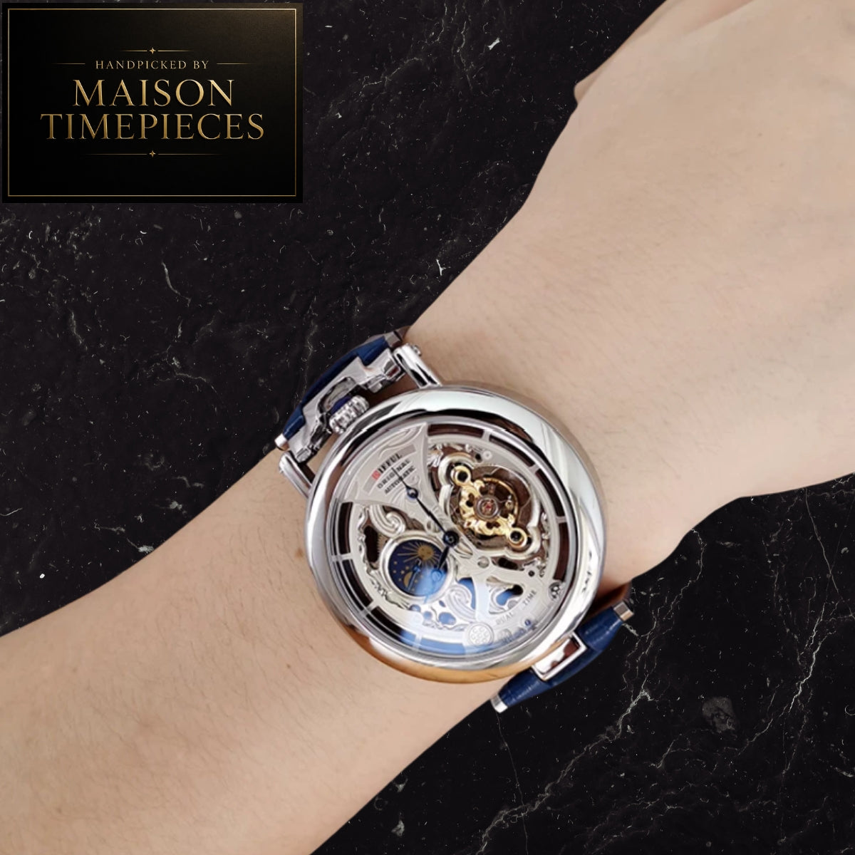 A close-up of a wrist wearing a Maison Timepieces skeleton watch with a silver-toned case, transparent dial showing internal gears, and a blue strap, set against a black marble background with a brand logo in the top left.