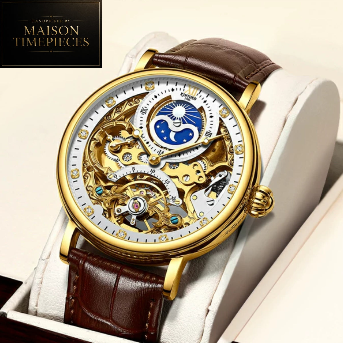 A luxury skeleton watch with a gold case, brown leather strap, and a detailed white dial showing the inner mechanical movement. The watch is displayed on a white cushion inside a box, with a 'Maison Timepieces' logo in the top-left corner.