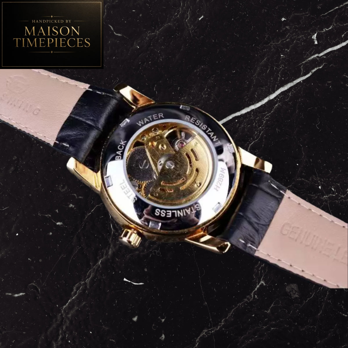 A close-up of a wristwatch shown from the back, revealing a gold-tone case, transparent crystal, and visible mechanical movement. The watch has a black leather strap with a light beige underside and white stitching.