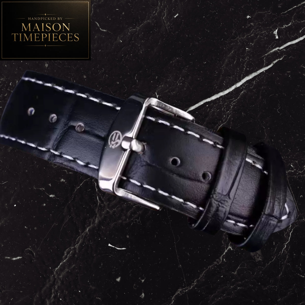 A close-up of a black leather watch strap with white contrast stitching and a silver-toned buckle, laid diagonally on a dark marble surface. A Maison Timepieces logo badge is visible in the top-left corner.