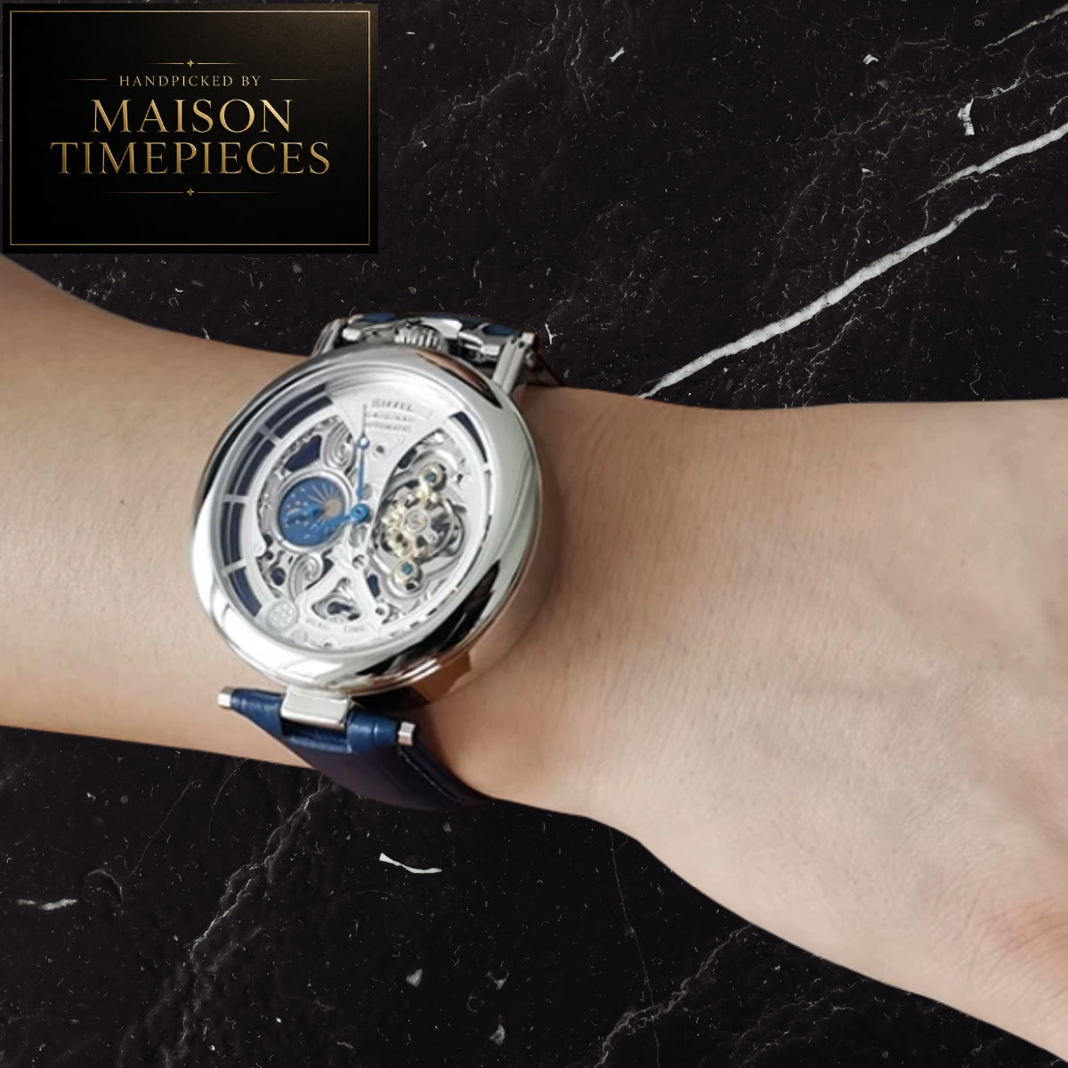 A close-up of a wrist wearing a Maison Timepieces skeleton watch with a silver-toned case, white dial, and blue strap, set against a black marble background with a Maison Timepieces logo in the top-left corner.