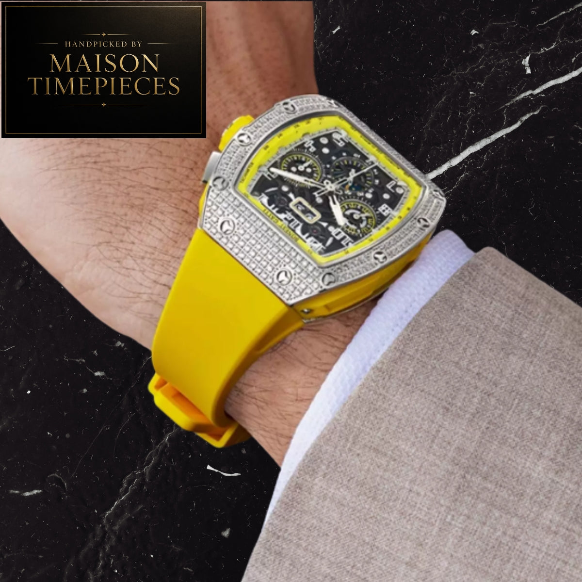 A close-up of a wrist wearing a Maison Timepieces luxury watch with a bright yellow silicone strap, a silver-tone bezel paved with clear crystals, and a black dial with yellow accents, set against a black marble background.