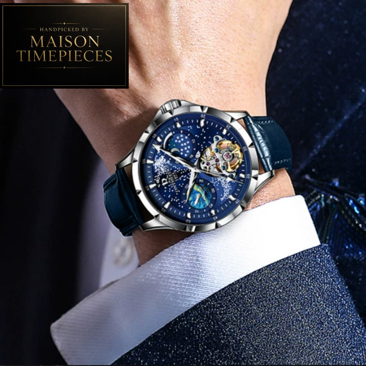 A close-up of a wrist wearing a Maison Timepieces skeleton watch with a deep blue starry dial, silver-tone case, and dark blue leather strap, shown against a dark suit and white shirt cuff.