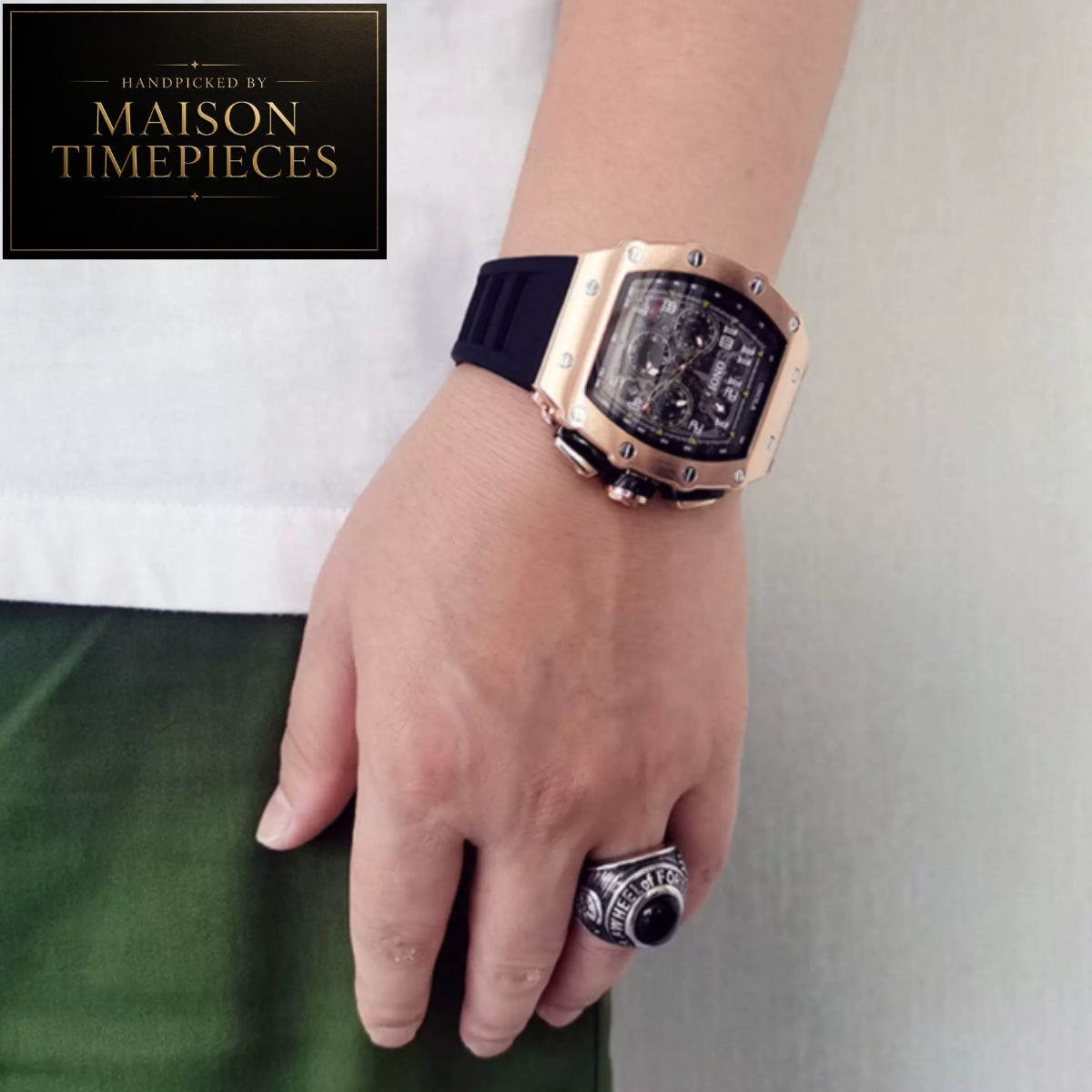 A close-up of a person's hand and wrist wearing a Maison Timepieces watch with a rose gold case, black dial, and black strap, paired with a silver ring. The background shows a white shirt and green pants.
