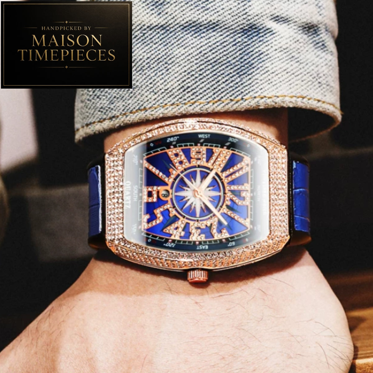 A luxury wristwatch with a rectangular rose gold case, a crystal-encrusted bezel, a blue dial with gold Roman numerals, and a blue strap, shown worn on a wrist with a denim cuff. A black logo overlay in the top-left corner reads 'Handcrafted by Maison Timepieces'.