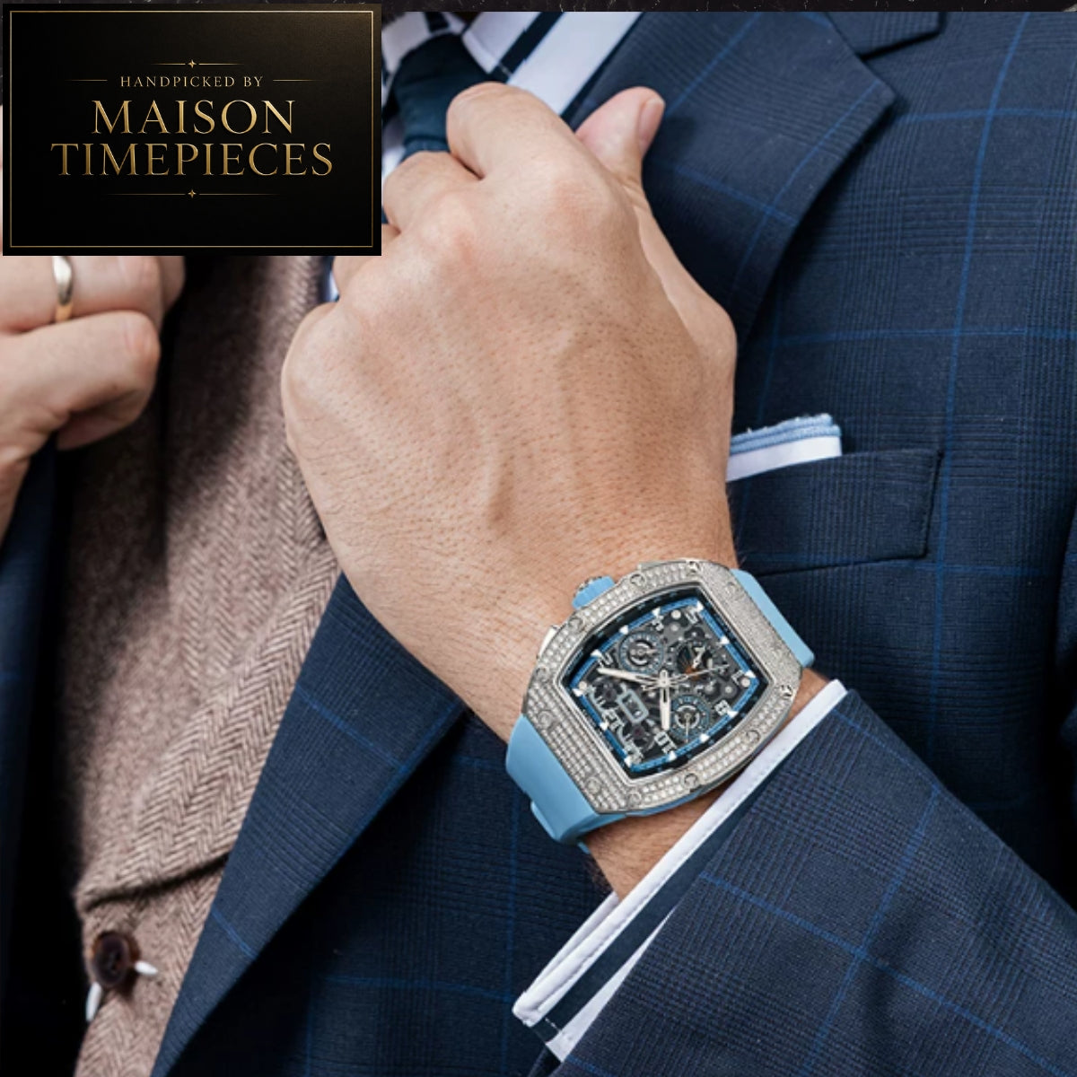 A luxury watch with a light blue strap and a diamond-encrusted bezel, shown on a man's wrist. The man is wearing a dark blue plaid suit jacket and a brown herringbone shirt.
