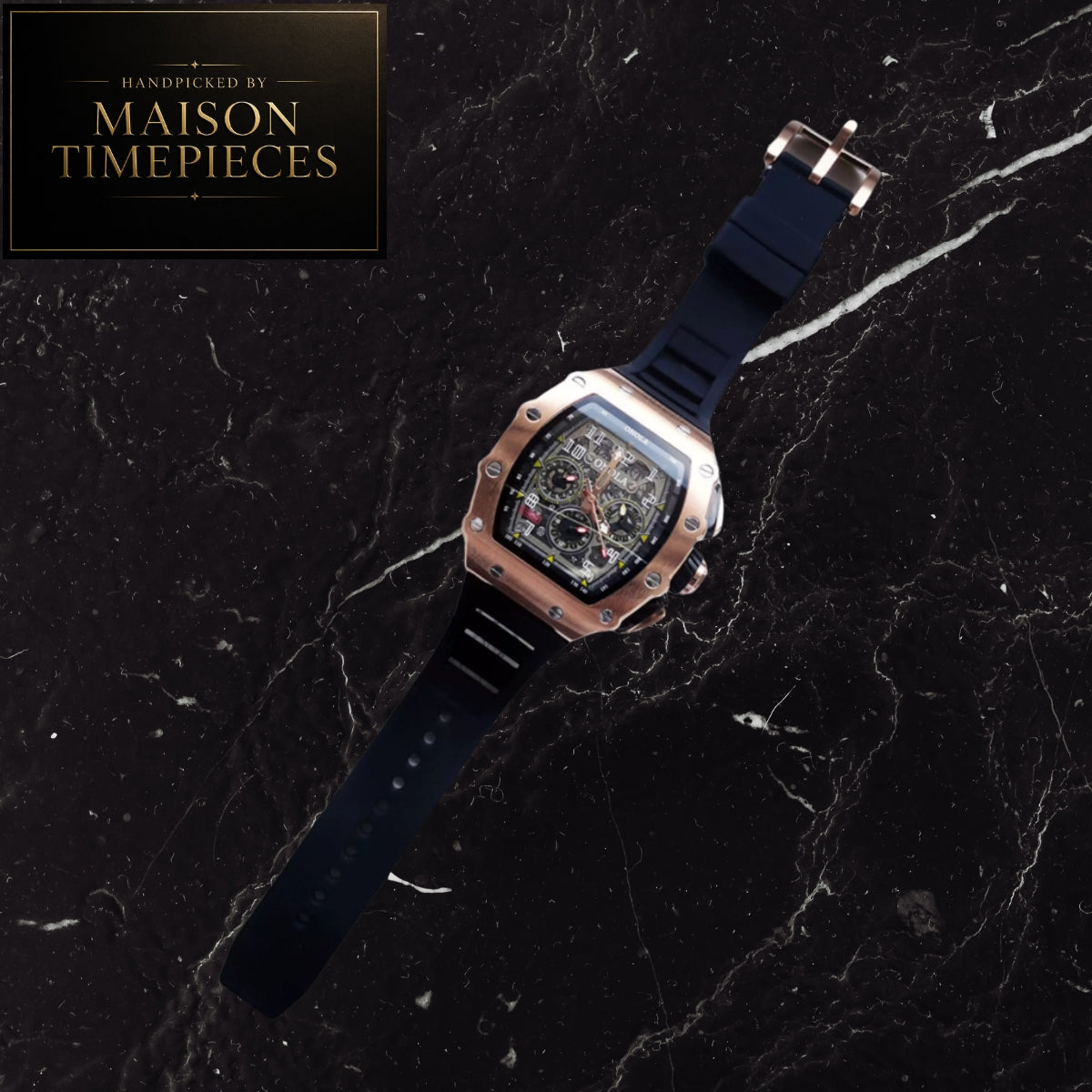 A luxury watch by Maison Timepieces with a rose gold-toned case, black dial, and dark navy blue strap, presented diagonally on a black marble background with a brand logo overlay in the top left corner.