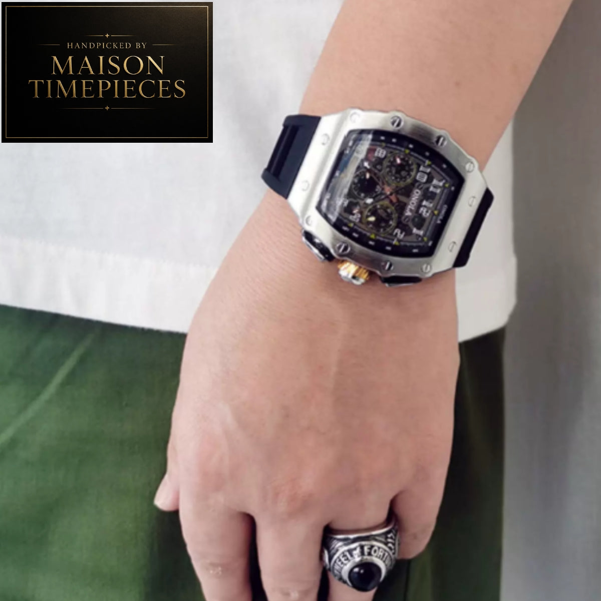 A close-up of a person's wrist wearing a Maison Timepieces skeleton watch with a silver-toned case, black dial, and black strap. The person is also wearing a silver ring, and a Maison Timepieces logo badge is visible in the top-left corner.