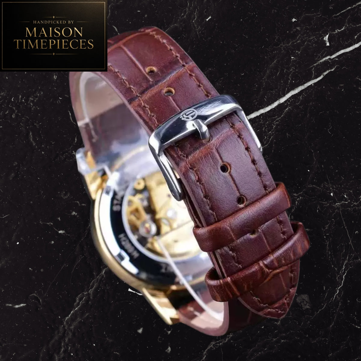 A close-up of a brown leather watch strap with a silver buckle, attached to a gold-toned watch case, photographed on a dark marble surface. A black square logo in the top-left corner reads 'Handpicked by Maison Timepieces'.