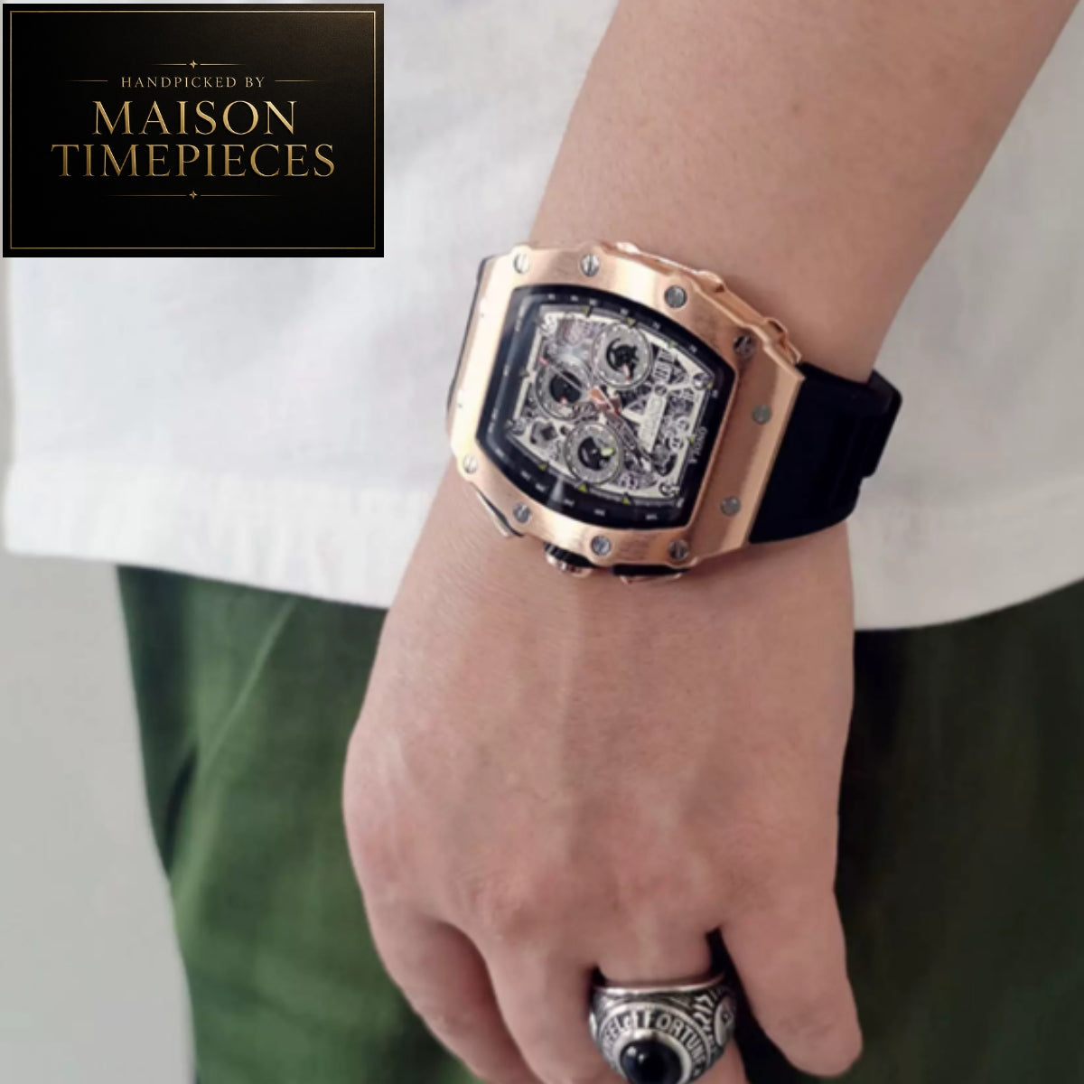 A close-up of a wrist wearing a Maison Timepieces watch with a rose gold-toned case, black strap, and skeleton dial, shown on a person's wrist with a white shirt and green pants in the background.