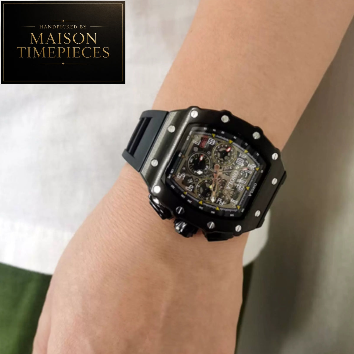 A close-up of a wrist wearing a Maison Timepieces skeleton watch with a black case, black strap, and transparent dial showing the inner mechanism. A Maison Timepieces logo badge is visible in the top-left corner.